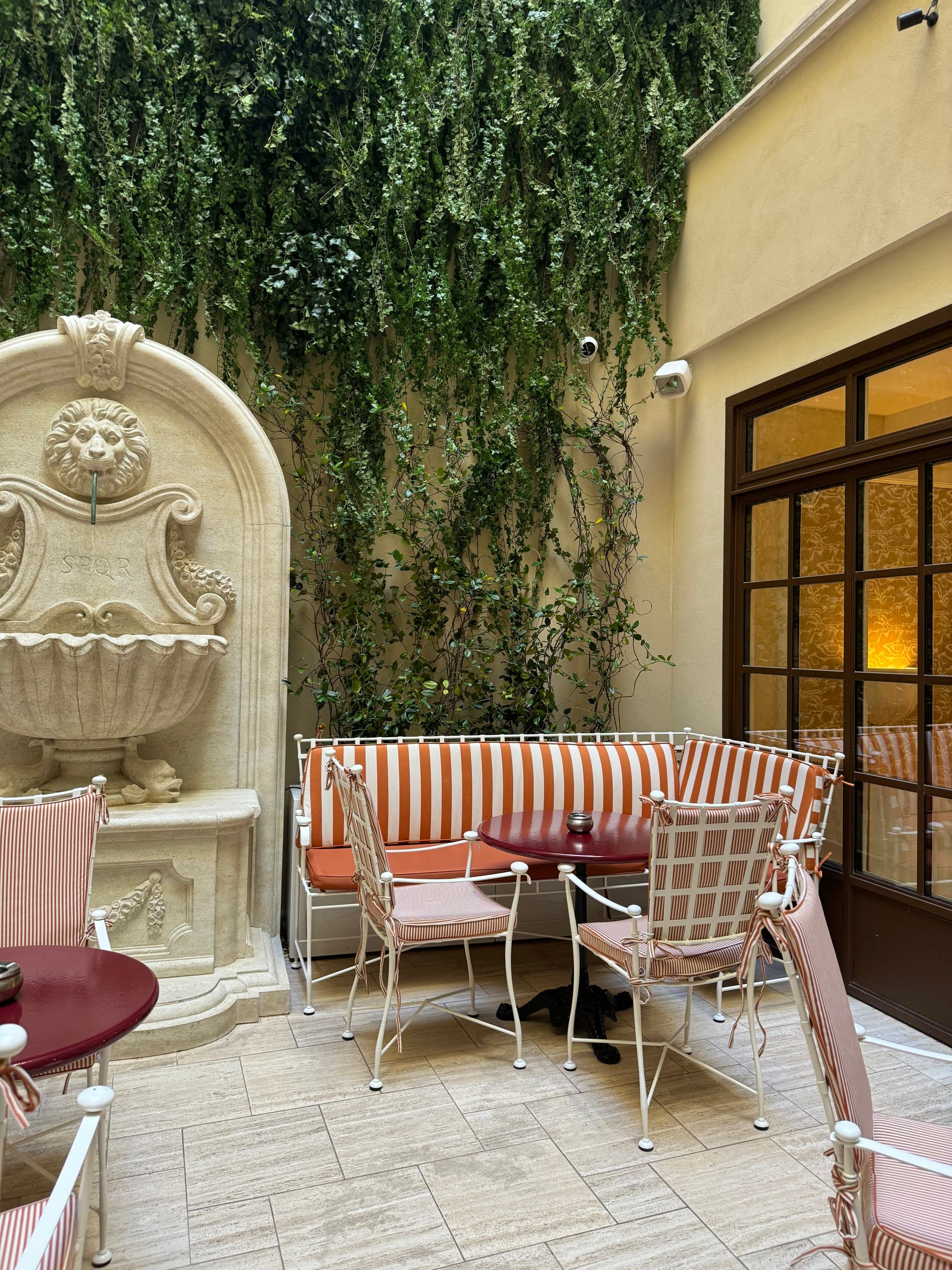 A view of outside seating at a hotel with a stone fountain, ivy covering one wall, a couch with red and white striped fabric and iron white chairs around a table.