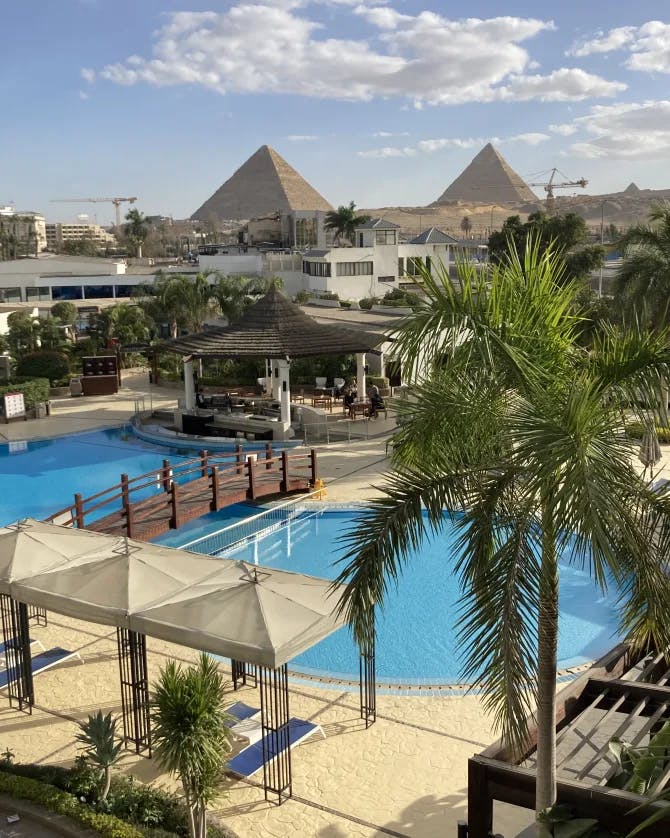 A resort with pyramids in the distance and a pool and palm trees.
