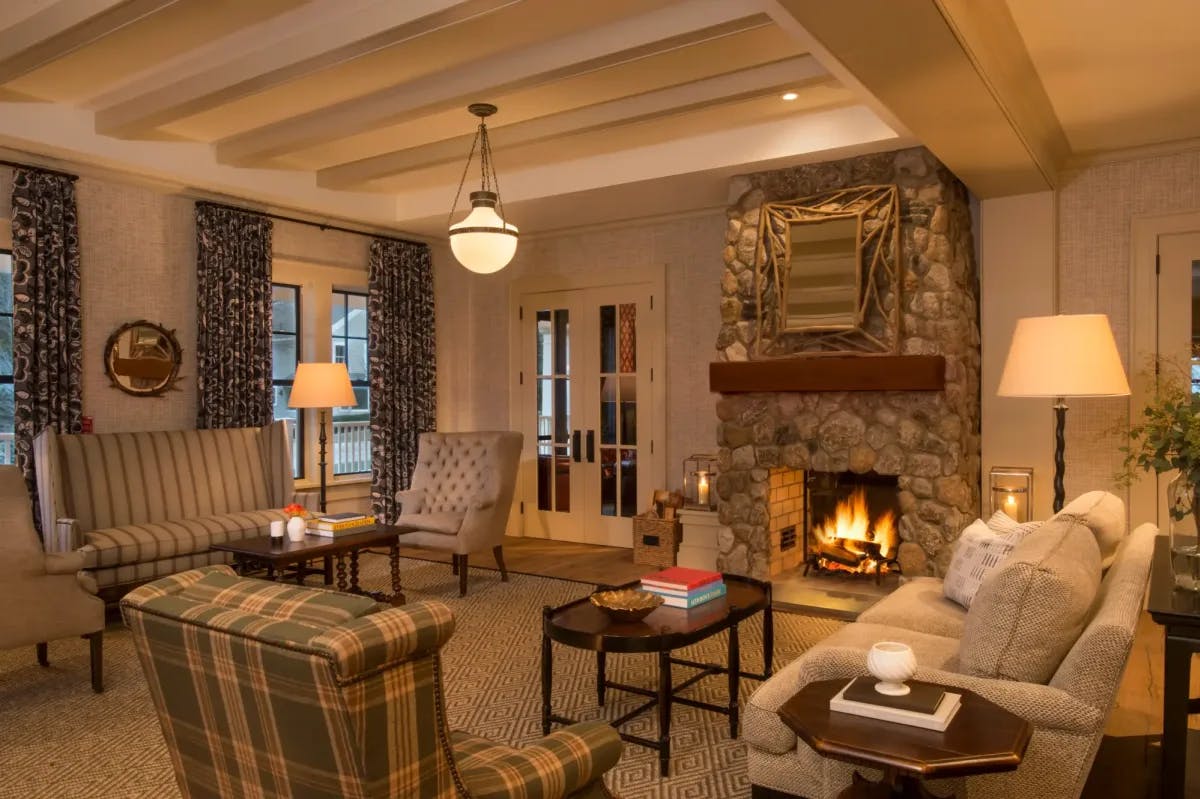 A cozy common area is lit by warm lighting and a stone fireplace. Classy furnishings fill the space, with fall gloom visible through shuttered windows.