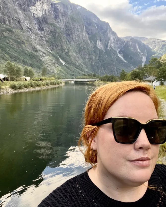 Emyli wearing a black top and sunglasses posing for a selfie in front of a lake and mountains