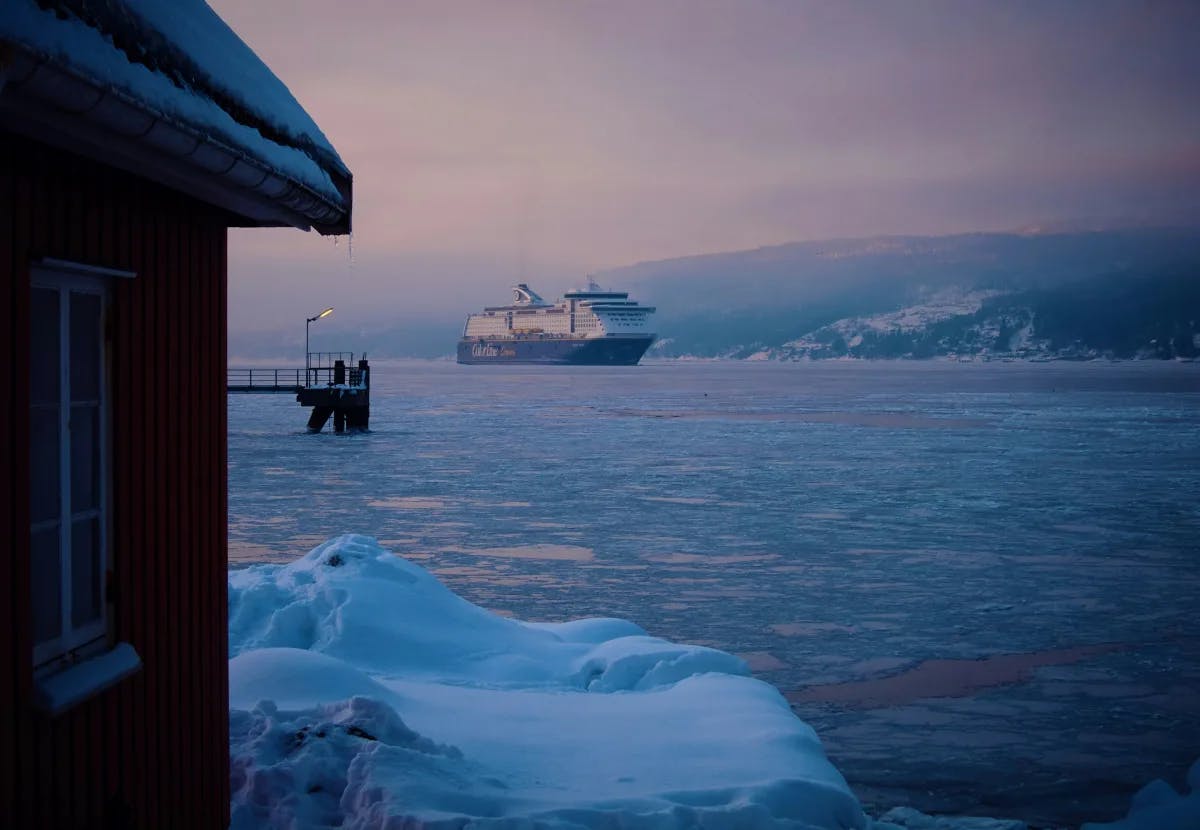 A side-view of a house, looking out onto the sea with an icebreaker ship, and snowy mountains in the distance underneath a lavender sunset.