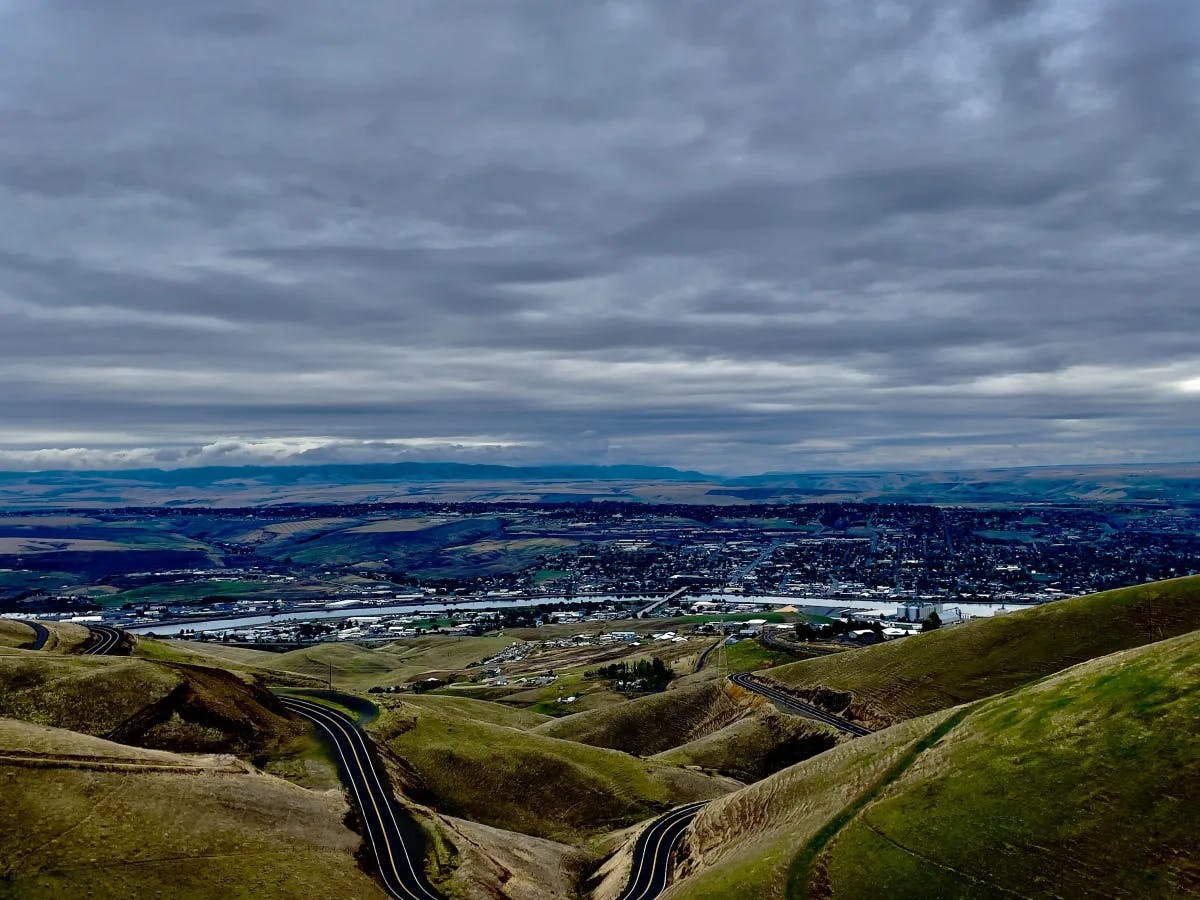 The image offers a panoramic view of a town cradled in a valley, with rolling hills under a dramatic cloudy sky.