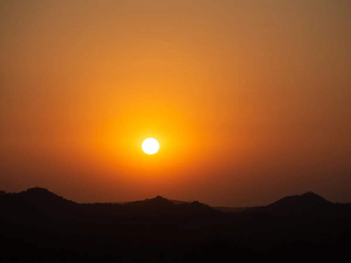 The image depicts a sunset with the sun prominently visible above silhouetted hills against an orange sky.