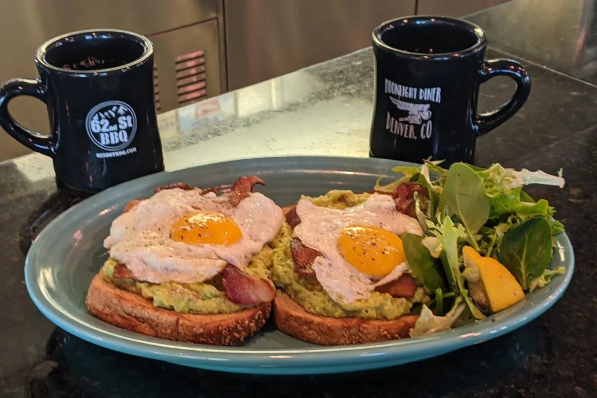 Eggs and bacon on avocado toast on a blue plate sitting in front of mugs