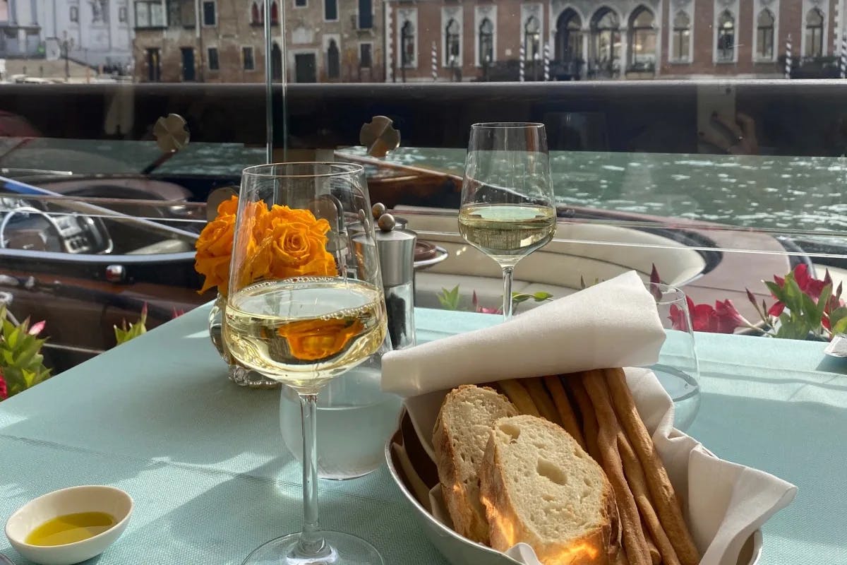 Outdoor dining setup with glasses of white wine and a bread basket on a table overlooking Venice canal.