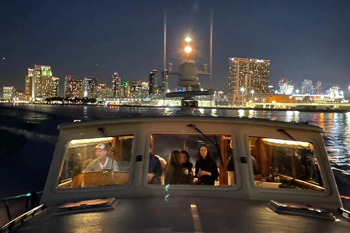 A picture of people inside of a yacht anchored in the harbor with a city skyline in the background lit up at night. 