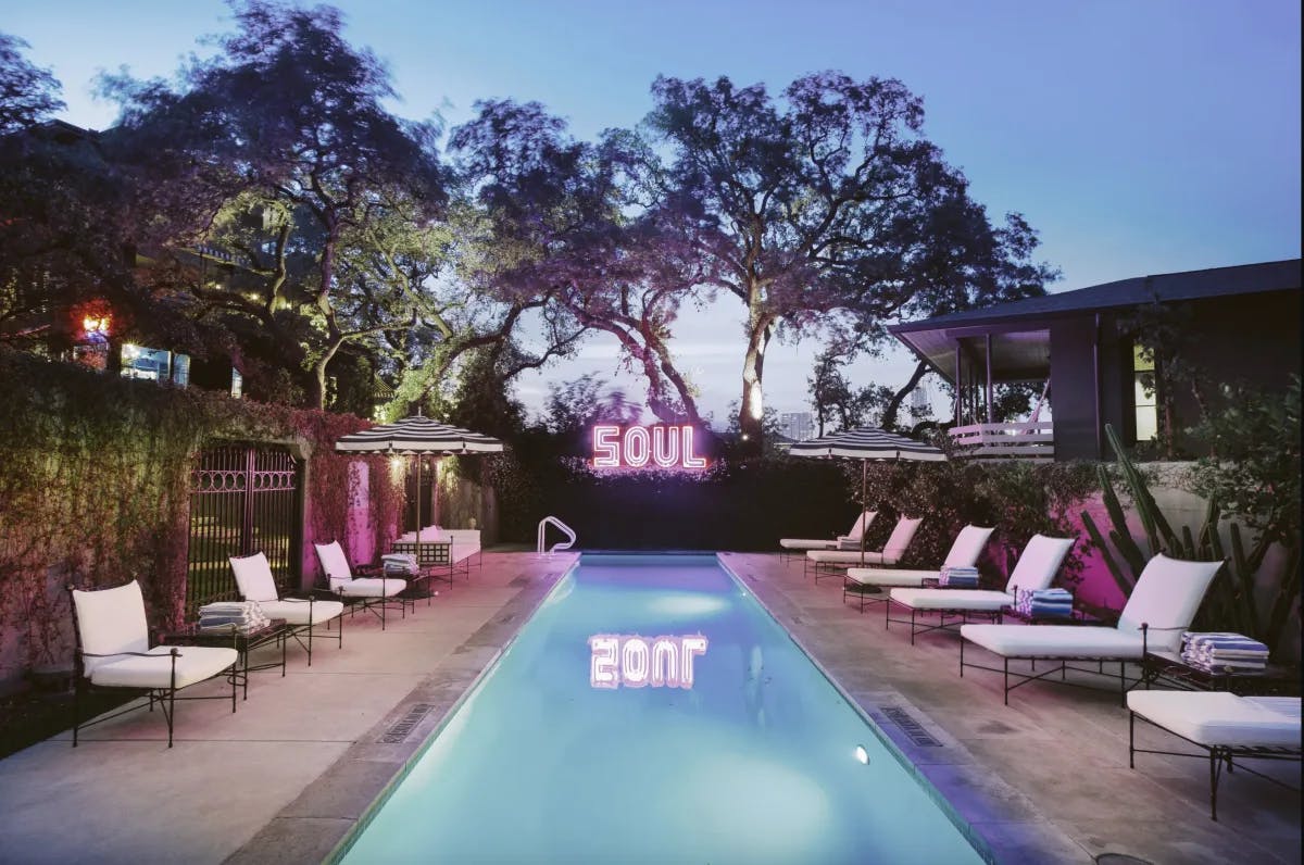 A swimming pool surrounded by white lounge chairs, plants, trees and a bright pink neon sign that says "SOUL". 