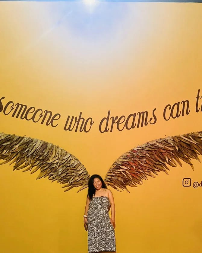 Rogelyn in a long dress posing against a wall mural that makes it look as if she has wings, reading “only someone who dreams can truly fly”