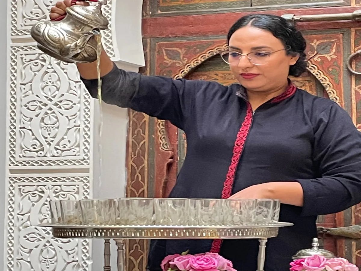 A picture of a woman holding metal teapot, pouring liquid into glasses on a table.