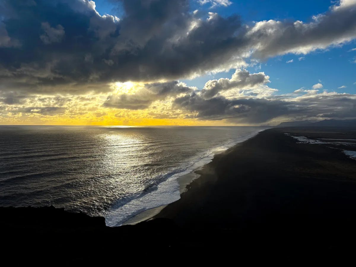 A view from off of Dyrholaey Cliff of a beach and water at sunset.