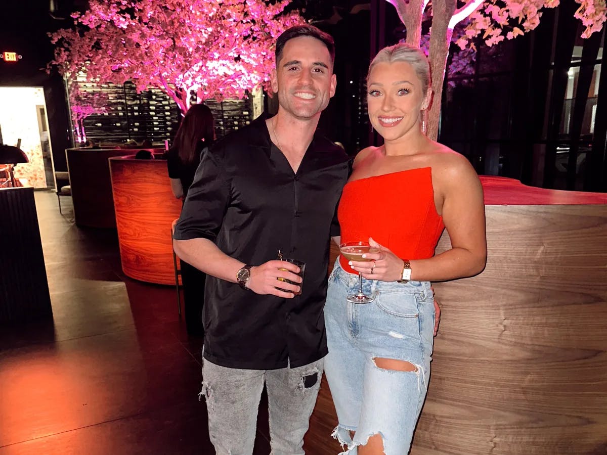 A woman in a red tank top and jeans and a man in a black shirt standing in front of a pink colored tree at a restaurant and holding drinks.