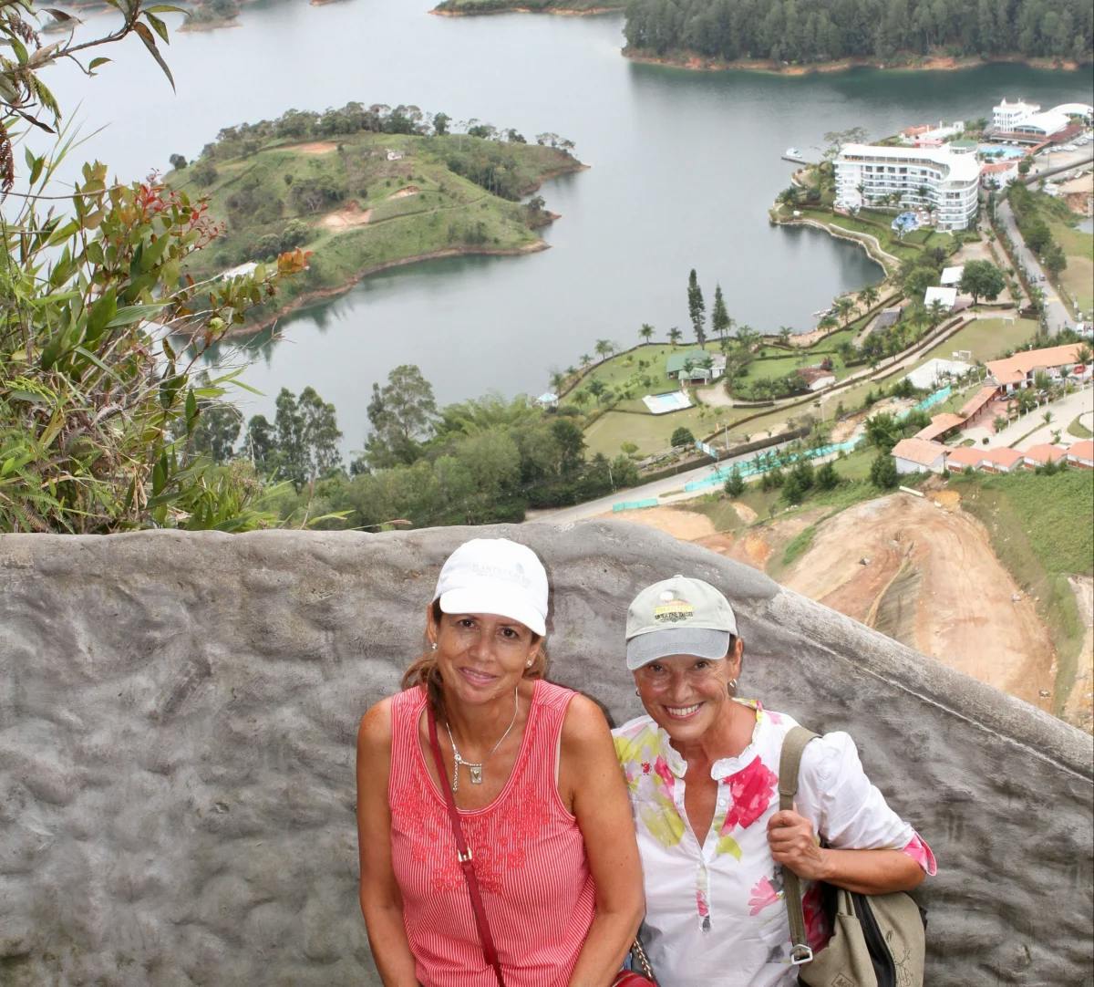 Two individuals stand atop a scenic overlook, gazing upon a landscape dotted with water and buildings, their faces veiled in mystery.