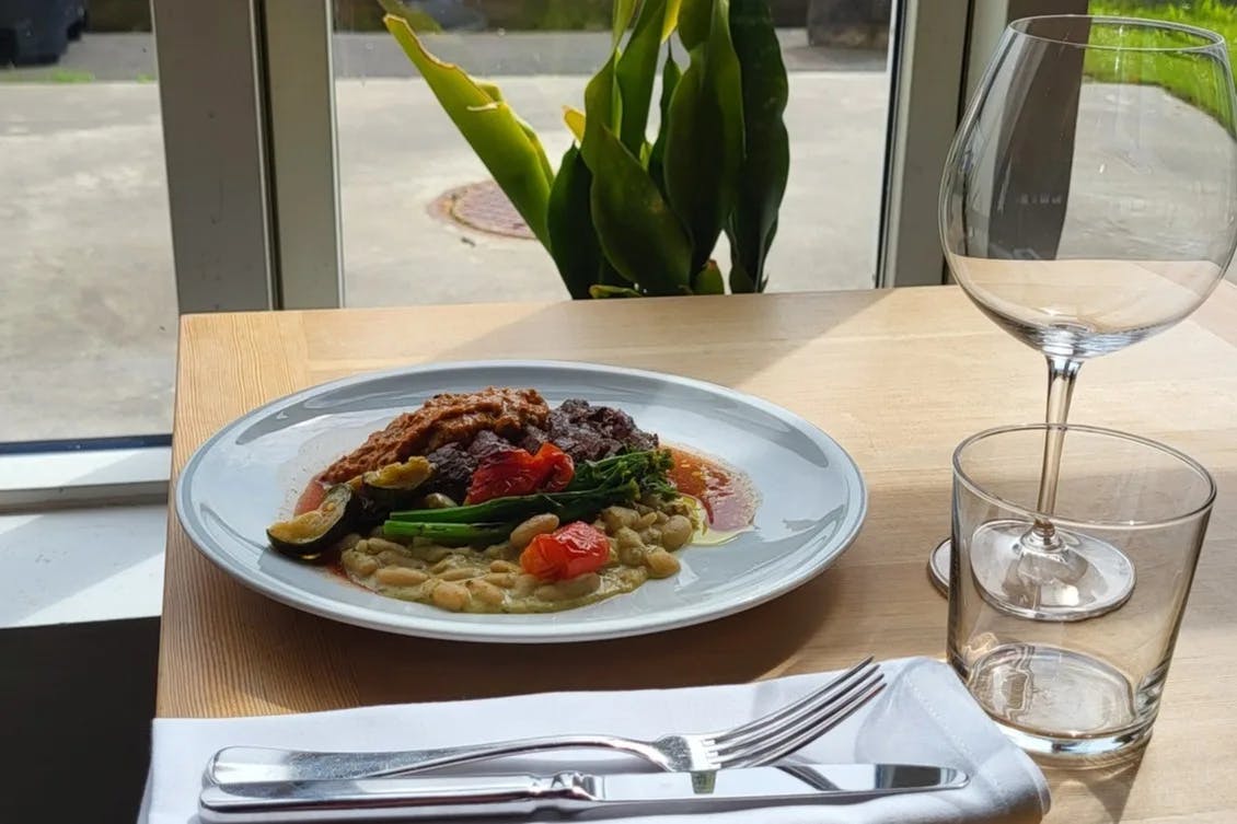 A plate of pasta and other colorful foods on top of a wooden table next to a wine and water glass, with a napkin and silverware in front of it. 