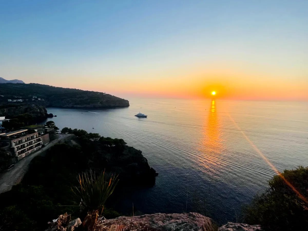 The image depicts a tranquil sunset over the sea, with a boat sailing towards the horizon and buildings nestled on the shore.