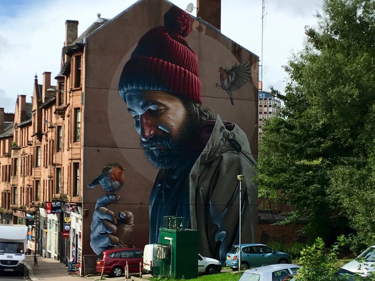 A mural of a man holding a bird on a building surrounded by trees and cars.