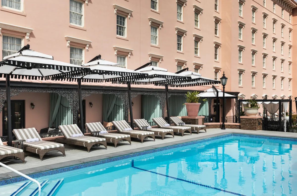 An outdoor swimming pool at a Charleston hotel, with striped lounge chairs, and white umbrellas with black fringe by the side of the pool.