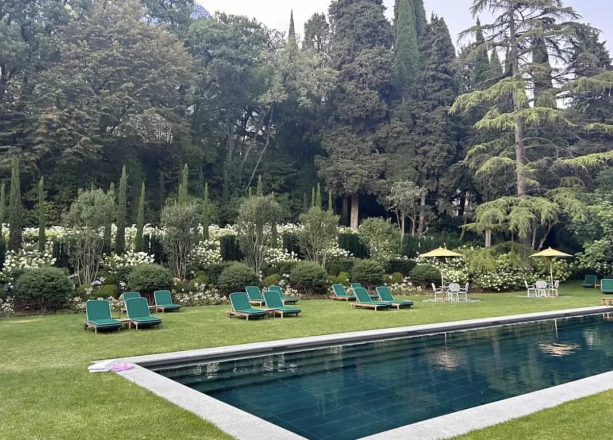 A stunning view of a swimming pool surrounded by green lawn chairs, tables, umbrellas, landscaping and gorgeous trees on top of a manicured grassy lawn. 