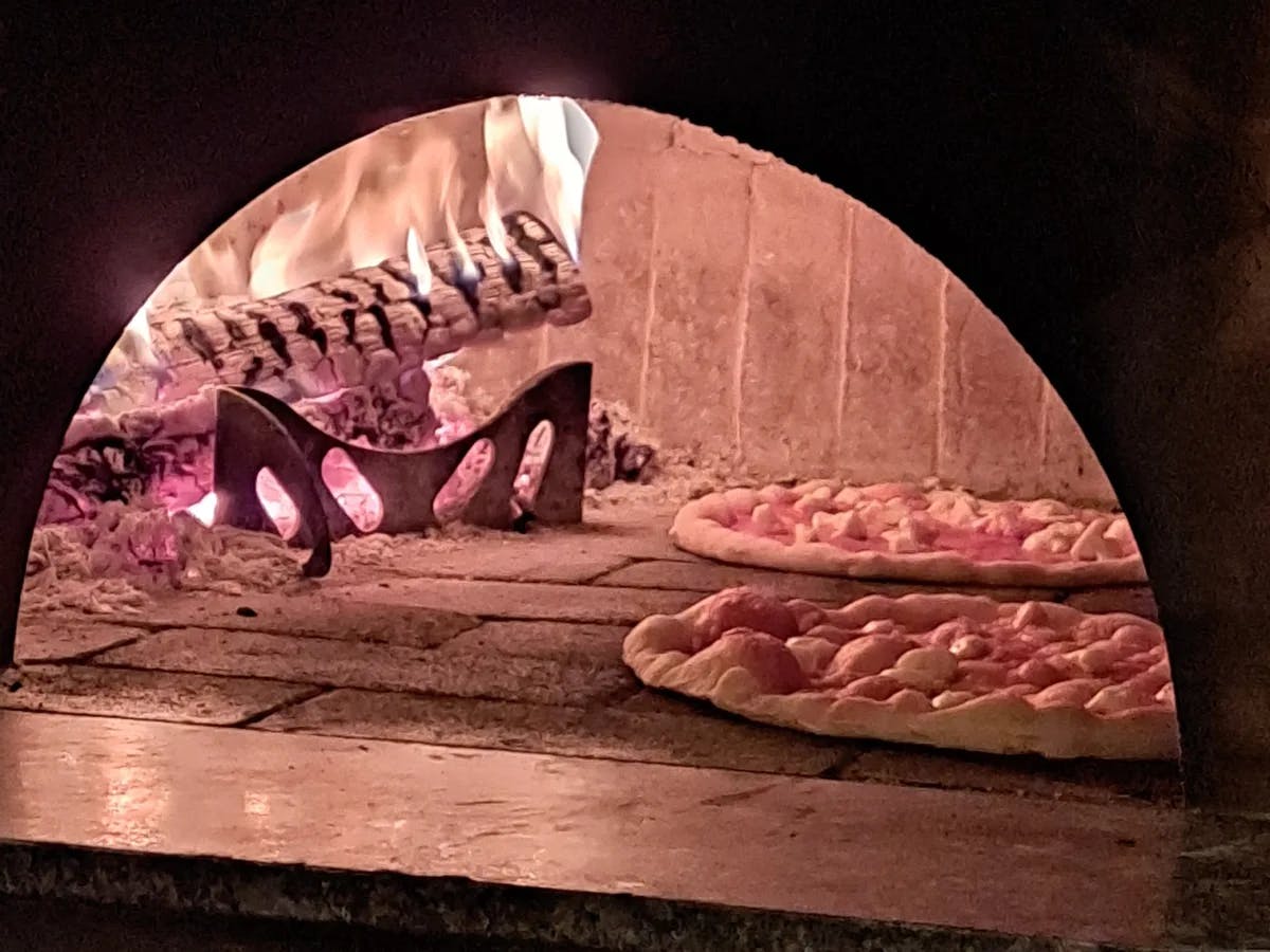 A view of two pizzas cooking inside of a pizza oven next to a log on fire. 