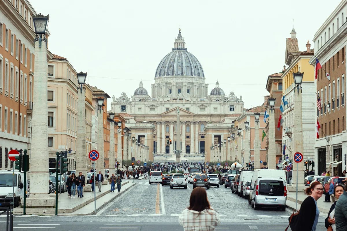 People walking towards St. Peter's Basilica, a large domed building between two streets.