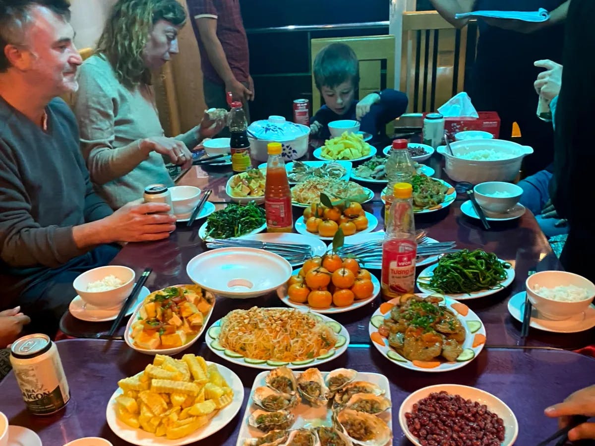 A table with various dishes and drinks is set, surrounded by people enjoying a communal meal.