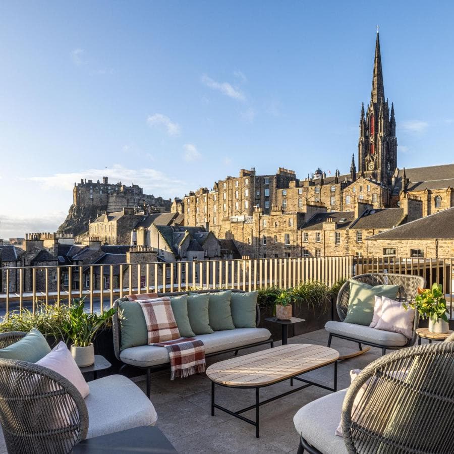 A view of an outdoor patio with couches, a table and a stone castle in the background surrounded by stone buildings and a church tower. 