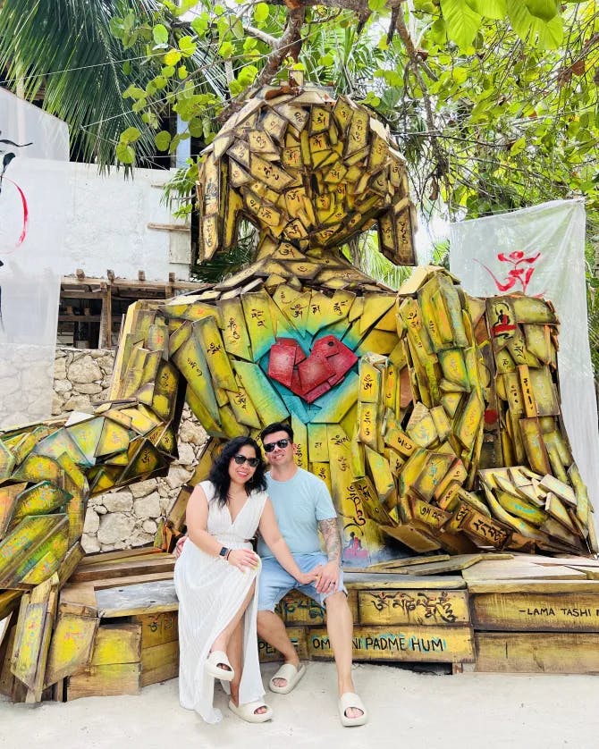 Rogelyn and a man posing for a photo in front of a yellow wooden sculpture shaped like a human