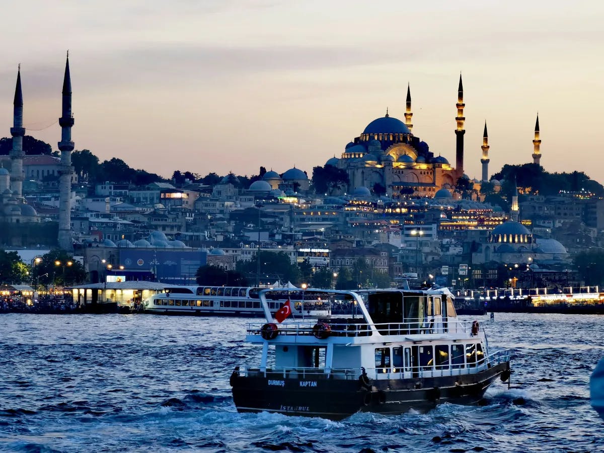 The image captures a scenic cityscape at dusk, featuring the silhouette of a mosque’s domes and minarets with a boat sailing in the foreground.