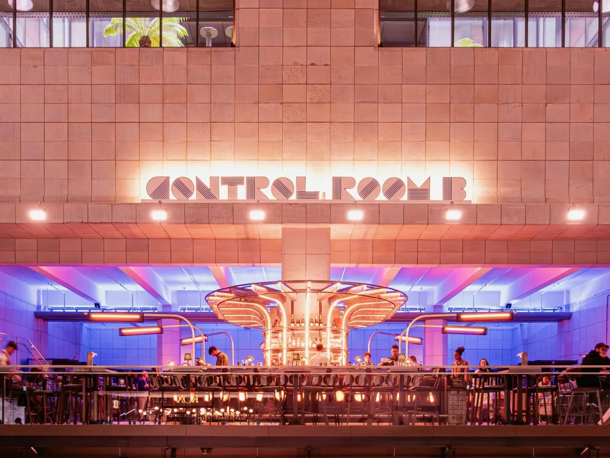 The image shows an interior space featuring a bar with a distinctive, illuminated central structure, surrounded by patrons, under a sign that reads “CONTROL ROOM B”.