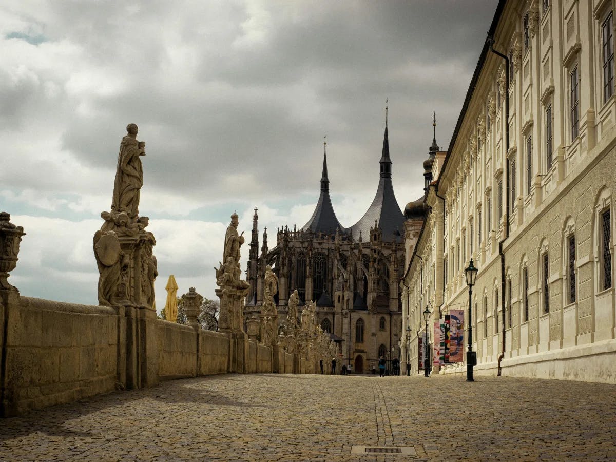 The image features a cobblestone street leading to a gothic cathedral, lined with statues under a cloudy sky.