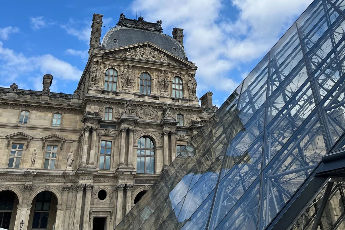 An exterior shot of the entrance of the Louvre museum, with a side of the glass pyramid in the front, and the stone building and its carved statues behind it.