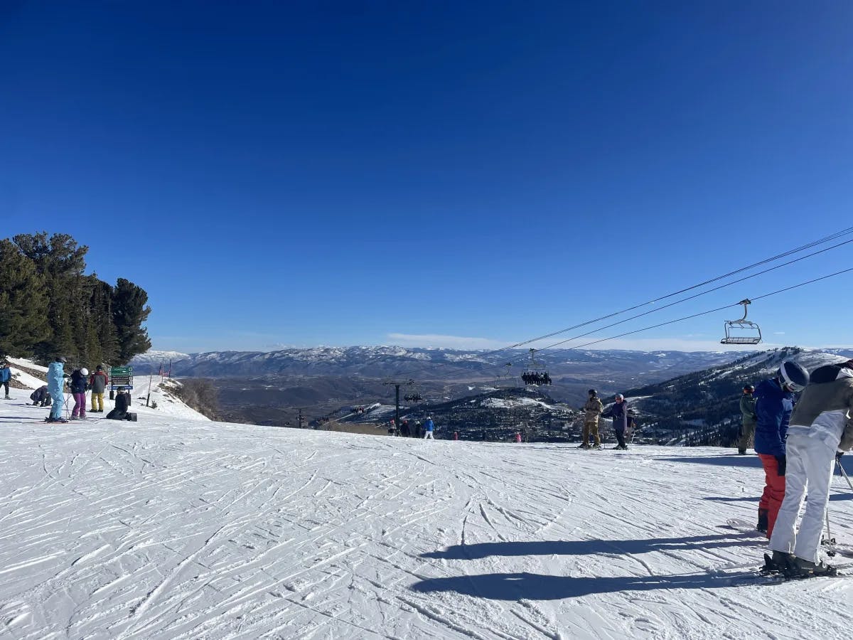 A snowy mountain top with various people wearing ski gear. There is a mountain range in the background and a ski lift to the right. 