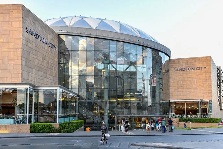 This image features a large building with brick walls and a geometric glass dome called Sandton City, it is a shopping and dining destination.