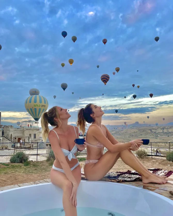 Camille and a friend posing in a jacuzzi overlooking a valley with several hot air balloons in the sky above