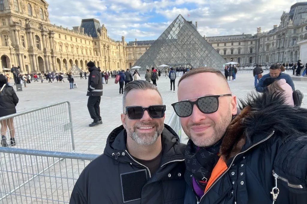 two men in winter coats smiling in front of the Louvre pyramid in Paris