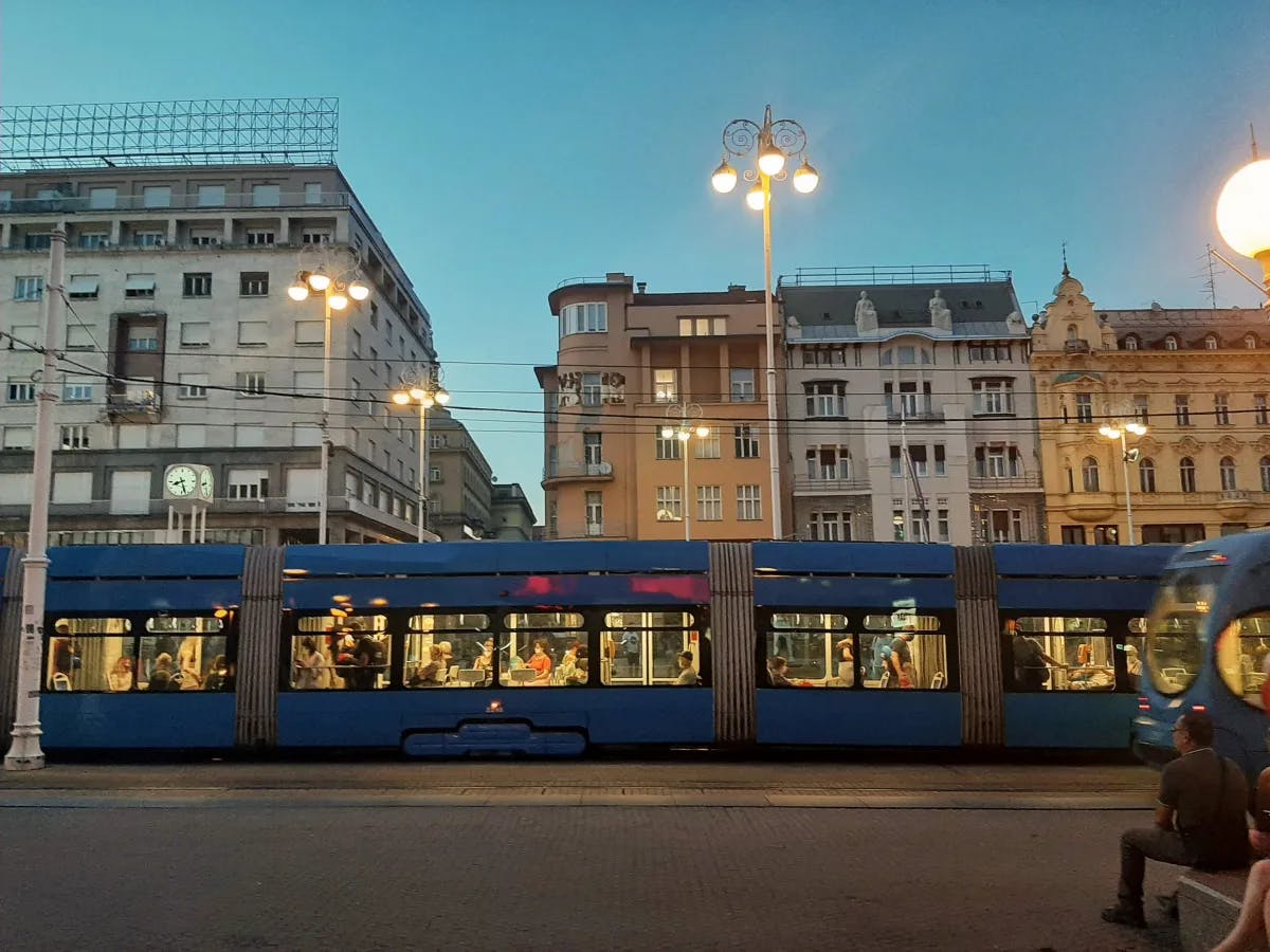 A blue train filled with passengers near the buildings during the evening time.