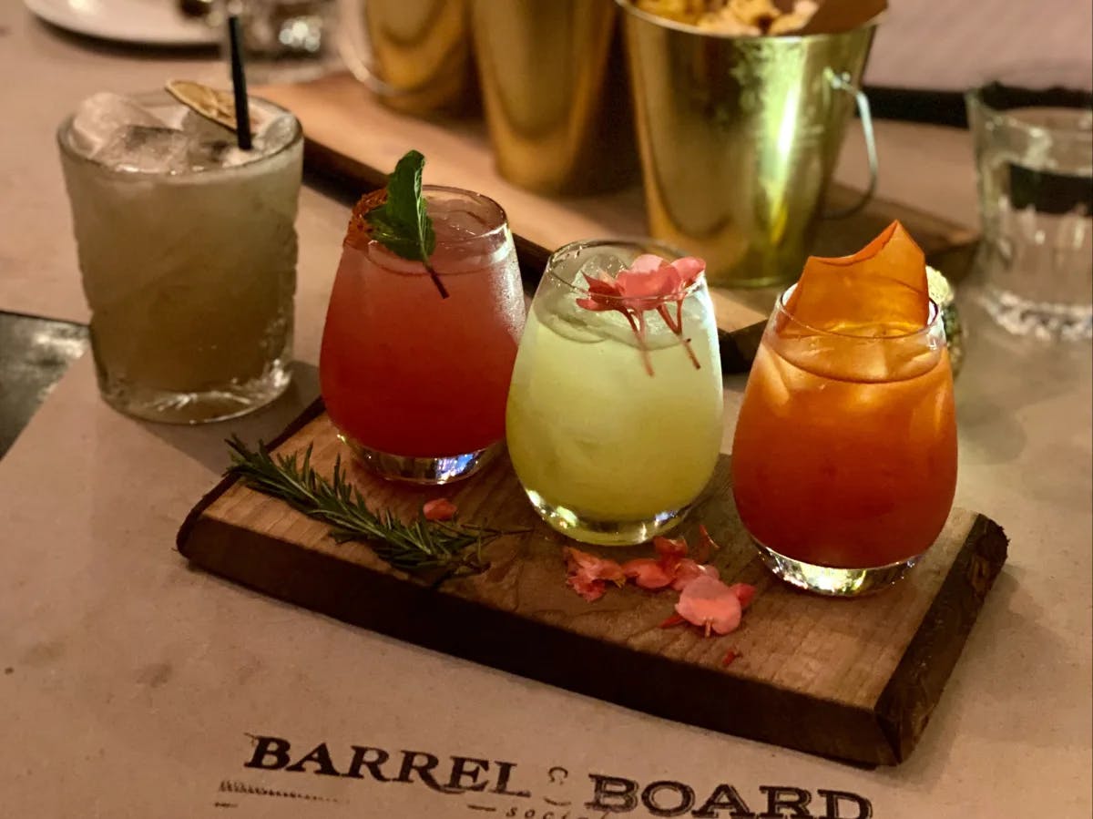 A wooden board with four different cocktails garnished with herbs and citrus is presented on a table with snacks in the background.