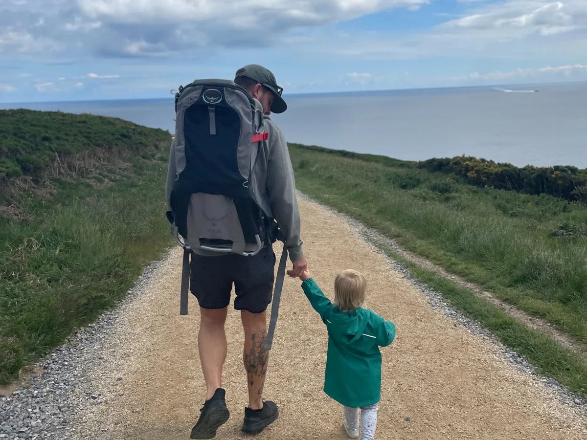 The image shows an adult walking hand-in-hand with a small child on a gravel path, surrounded by greenery and a distant sea view.