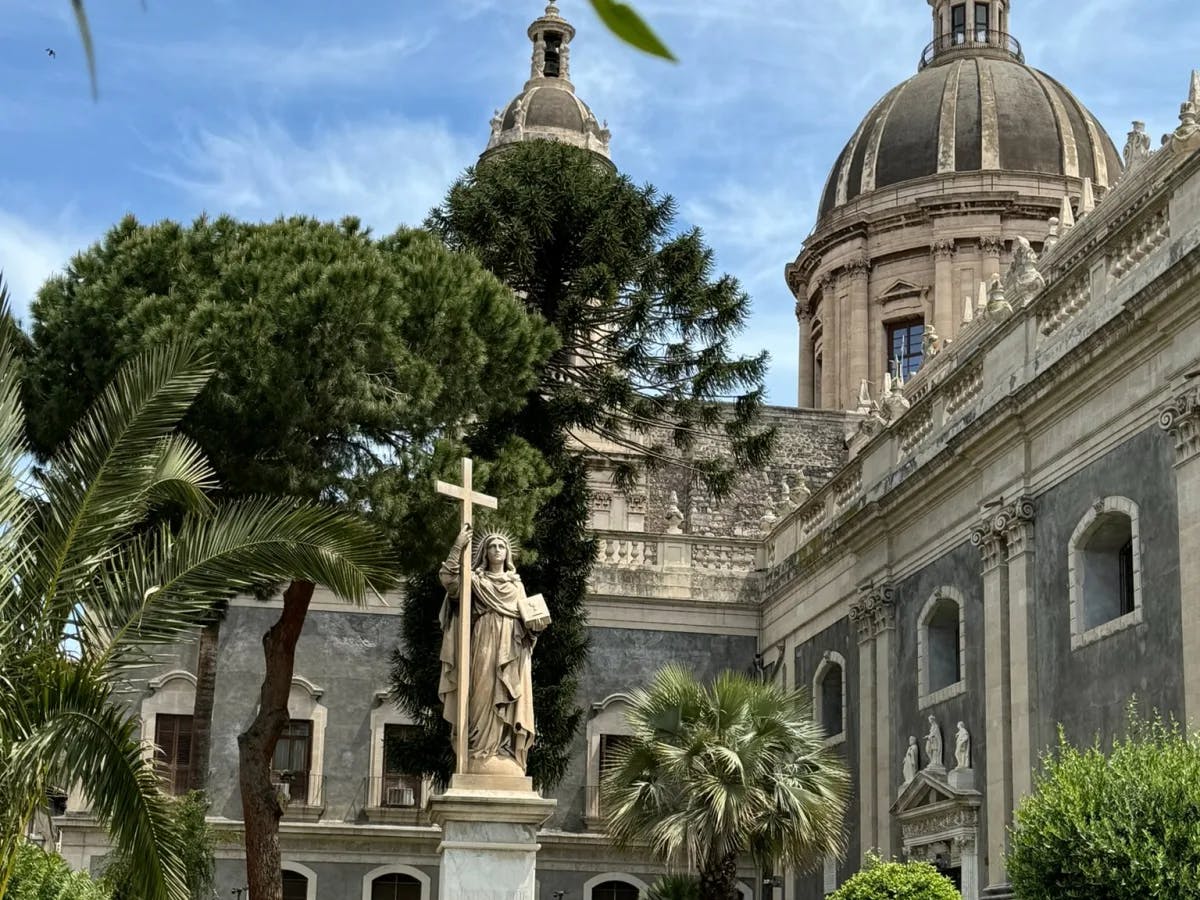 Cathedral building with domes and a statue of a woman holding a cross in its yard.