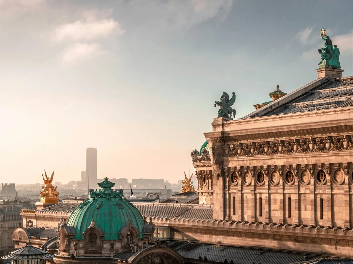 The image showcases a classical cityscape bathed in golden sunlight, highlighting a golden statue atop a grand building against a clear sky.