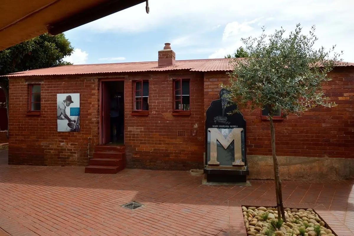 A small red brick building, with a large "M" and photo of Nelson Mandela on the outside.  The Mandela House, the former residence of Nelson Mandela, showcasing his life and legacy.