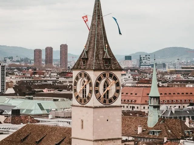 A stone and brown brick clock tower with a cityscape and mountain range in the background. 