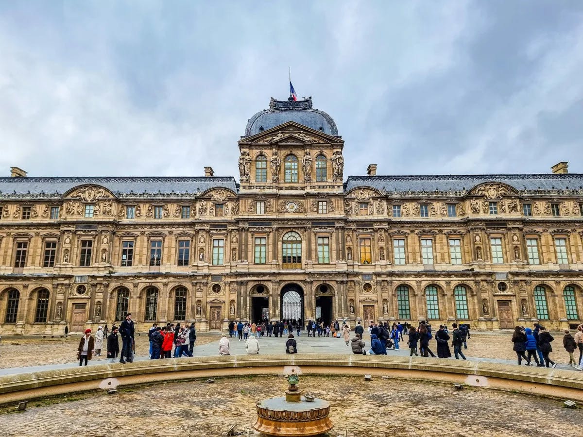 The image presents a grand historical building bustling with visitors in its courtyard, under a gloomy sky.