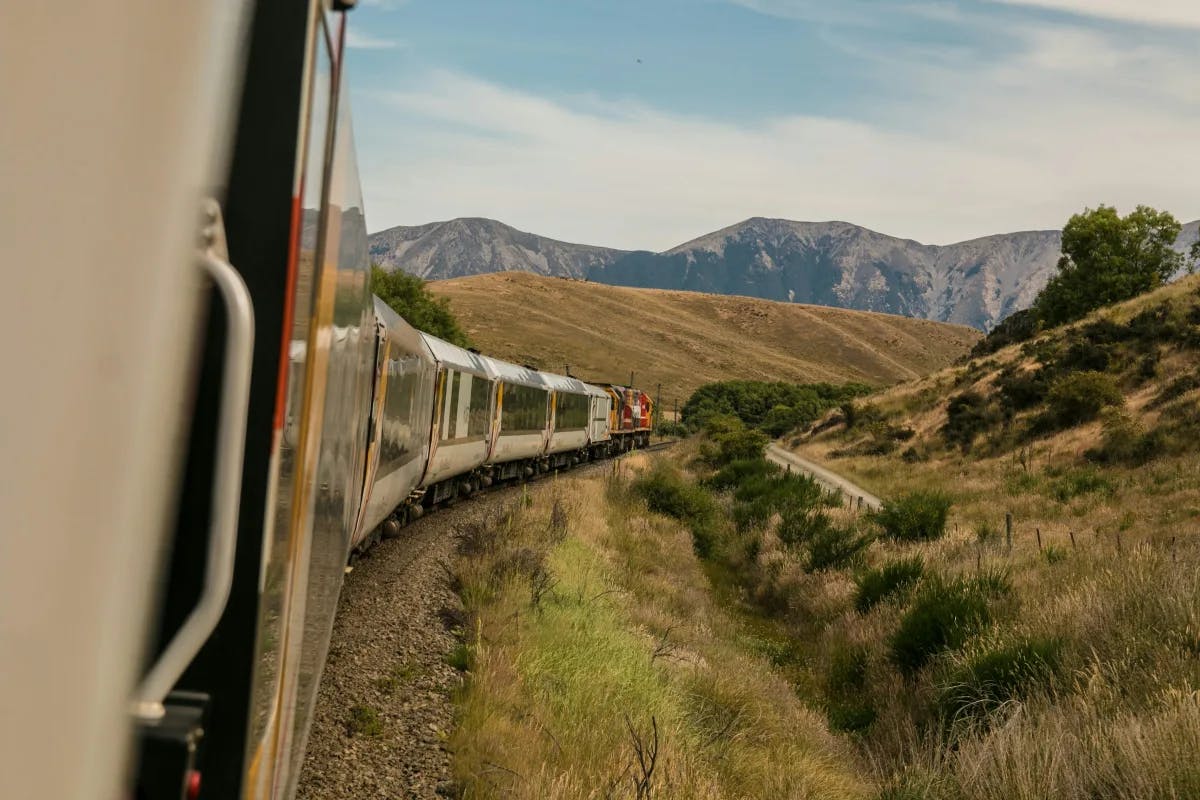 An image of a railway system going down a track with gorgeous views of rocky mountains and rolling green hills, shrubs and trees in the surrounding area. 