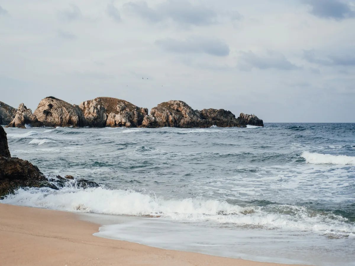 A tranquil beach scene with approaching waves and distant rocky outcrops under a cloudy sky.