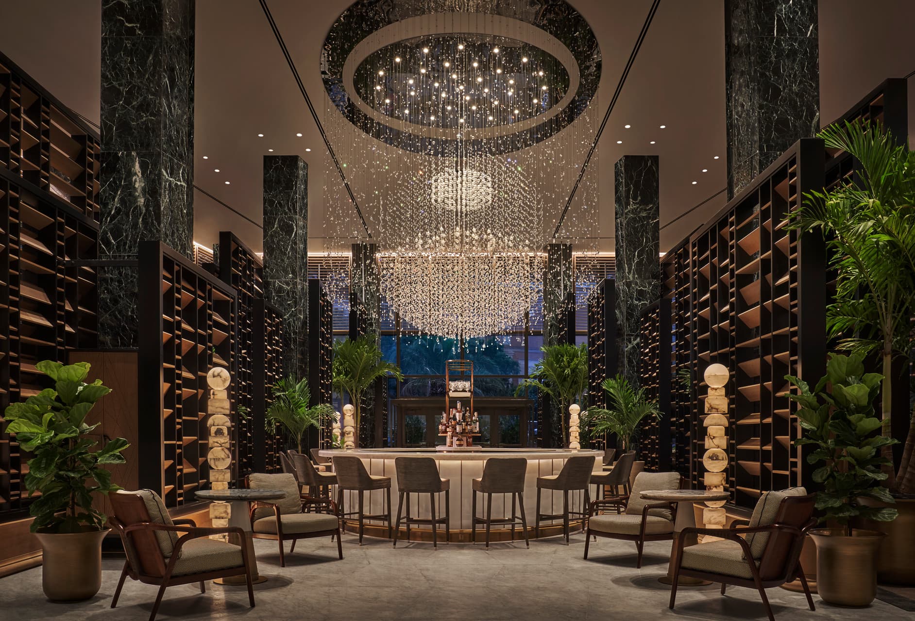 A view of a luxury hotel interior that is dimly lit with large pillars on either side, slatted wood walls, a circular table and chairs surrounding it with a large oval chandelier hanging from the ceiling.