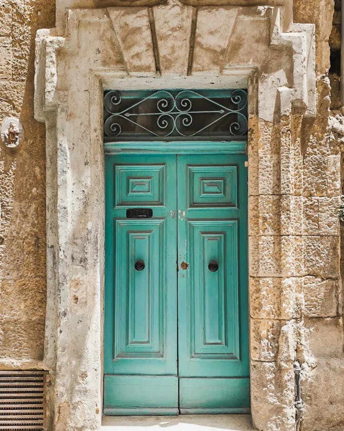A turquoise set of french doors with rustic hardware and a sand colored building with geometric designs. 