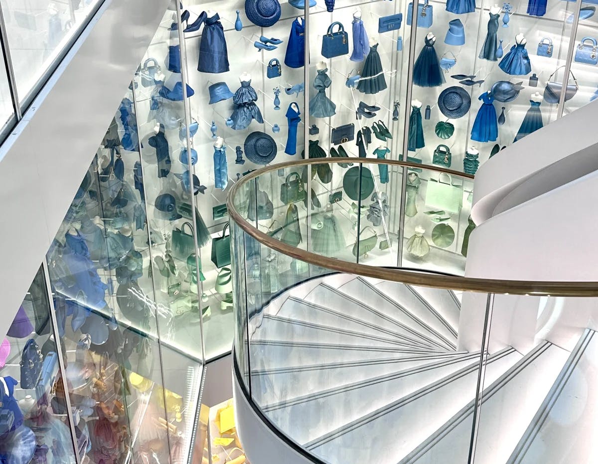 A white circular staircase with rainbow colored fashion pieces hanging behind multiple stories of glass at the Dior museum.