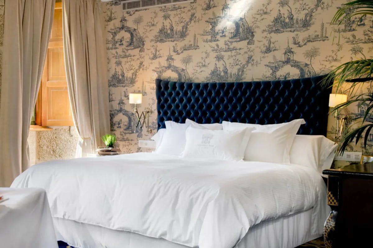 a bed with a blue velvet headboard in front of patterned wallpaper