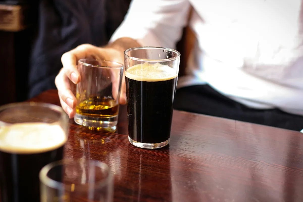 A wooden bar with a person's hand holding a shot of Irish whiskey next to two pint glasses of dark beer.