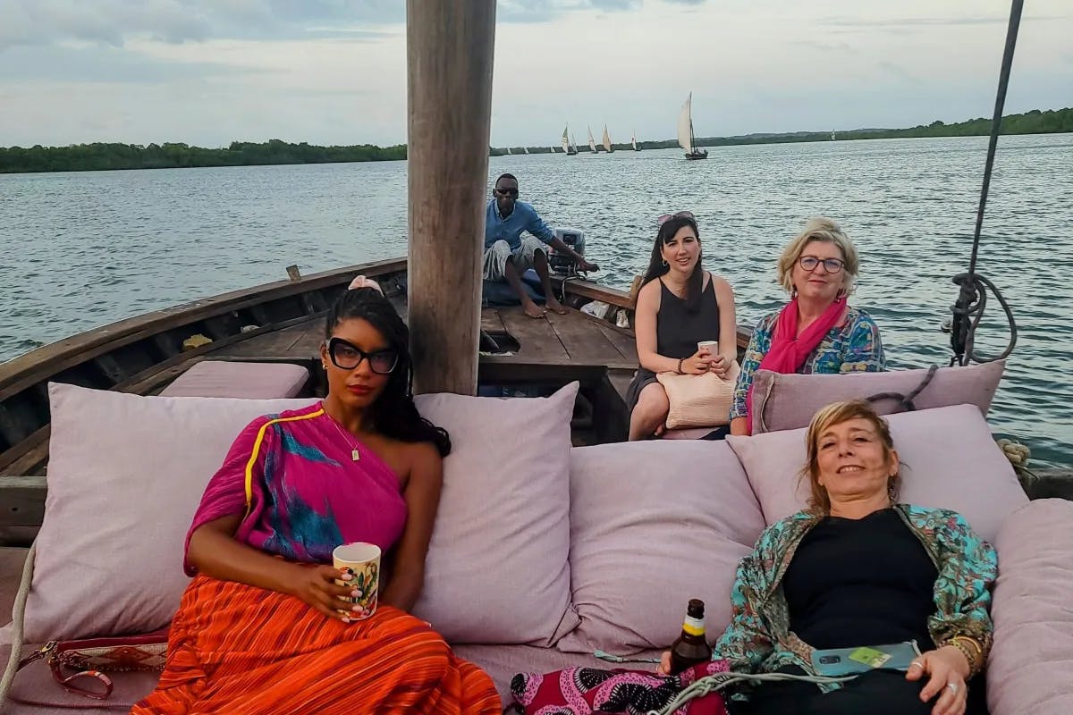 A view of five people lounging on a boat against large pillows. There is rippling blue water and other sail boats in the distance. 